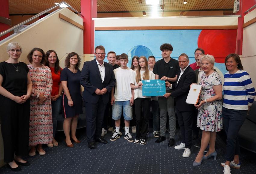 Das Gruppenfoto zeigt Verbraucherschutzminister Thorsten Glauber, Schüller und Lehrkräfte. Sie zeigen das überreichte Türschild Partnerschule und die Urkunde in die Kamera.