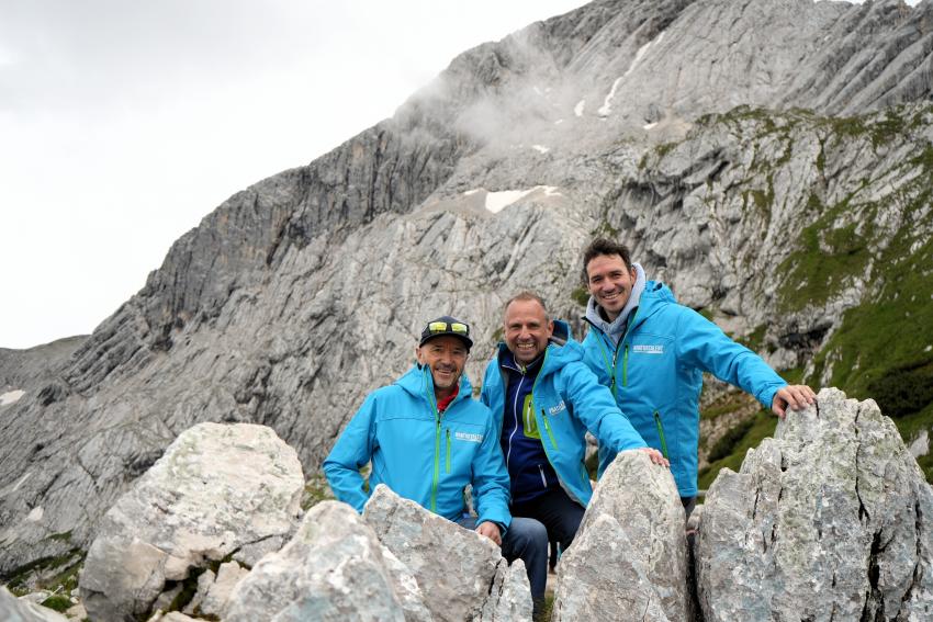 Umweltminister Thorsten Glauber (mitte), Christian Neureuther (links) und Felix Neureuther (rechts) stehen hinter Felsbrocken am neu eröffneten alpinen Themenweg 