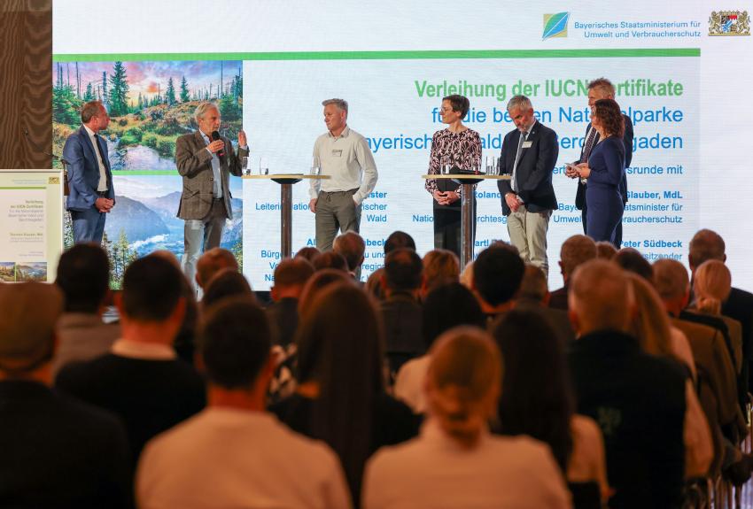 Auf der Bühne stehen von links nach rechts: Umweltminister Thorsten Glauber, Vorsitzender Zweckverband Bergerlebnis Berchtesgaden Dr. Bartl Wimmer spricht ins Mikrofon, Bürgermeister der Gemeinde Neuschönau Alfons Schinabeck, Leiterin des Nationalparks Bayerischer Wald Ursula Schuster (Foto links), Leiter des Nationalparks Berchtesgaden Dr. Roland Baier und Peter Südbeck, Vorstandsvorsitzender bei Nationale Naturlandschaften e. V.